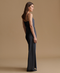 A woman modeling a floor-length dark gray satin slip dress with thin spaghetti straps, shown from a side-back profile against a solid warm beige studio background.