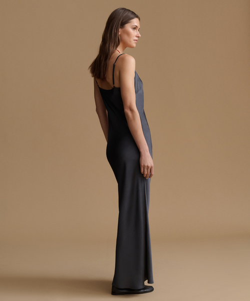 A woman modeling a floor-length dark gray satin slip dress with thin spaghetti straps, shown from a side-back profile against a solid warm beige studio background.