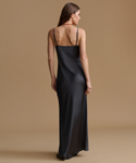 A woman wearing a black satin slip dress with thin straps, shown from the back against a solid beige studio background.