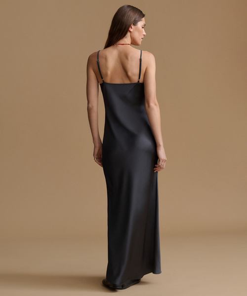 A woman wearing a black satin slip dress with thin straps, shown from the back against a solid beige studio background.