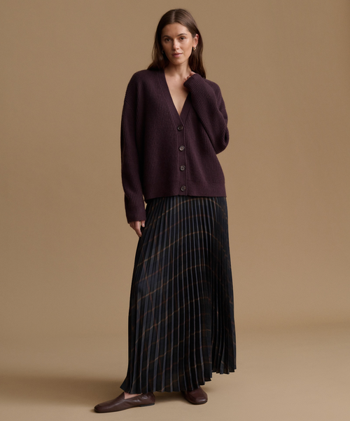 A woman models a dark plum cardigan sweater paired with a dark pleated skirt, standing against a solid beige studio background.