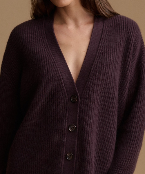 A close-up of a woman wearing a dark plum purple ribbed knit cardigan sweater with dark buttons, set against a solid warm beige background.