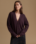 A woman wearing a dark plum ribbed knit cardigan with a deep V-neck and front buttons, paired with a dark pleated skirt, set against a solid warm beige studio background.