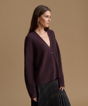 A woman wearing a dark plum ribbed cardigan with a V-neck and dark buttons, paired with a dark pleated skirt, photographed against a solid warm tan studio background.