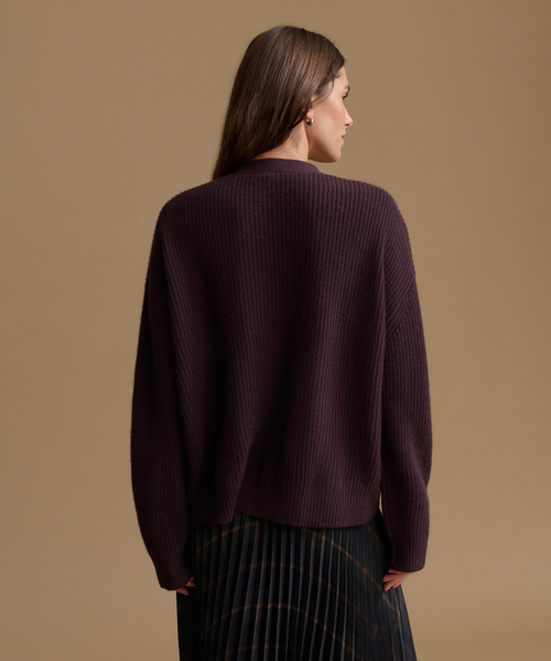 A woman shown from the back wearing a dark plum ribbed knit sweater and a dark pleated skirt, set against a solid warm beige studio background.