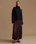 A woman stands against a plain warm beige studio background wearing a black ribbed turtleneck sweater and a dark burgundy pleated maxi skirt with black flats.