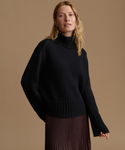 A woman wearing a black ribbed turtleneck sweater with wide sleeves and a pleated dark brown skirt, posed against a solid warm tan studio background.