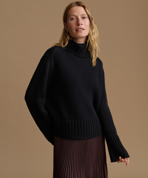 A woman wearing a black ribbed turtleneck sweater with wide sleeves and a pleated dark brown skirt, posed against a solid warm tan studio background.