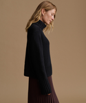 A woman in side profile wearing a black ribbed mock-neck knit sweater and a dark pleated skirt, set against a solid warm beige studio background.