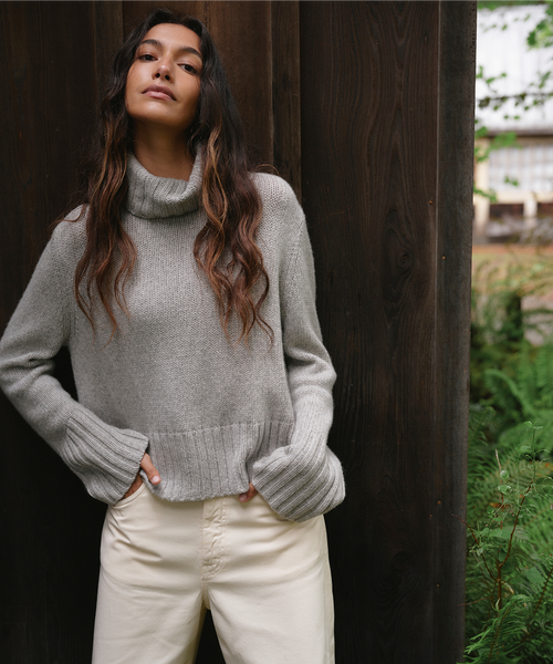 A woman wearing a light gray ribbed turtleneck sweater and cream-colored pants, standing against a dark wooden post with green foliage in the background.