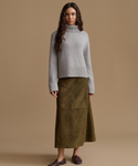 A woman modeling a light gray ribbed turtleneck sweater and an olive green midi skirt, standing against a solid tan studio background.