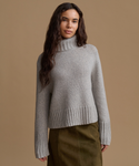 A woman wearing a light gray ribbed turtleneck sweater and an olive green skirt, standing against a solid tan studio background.