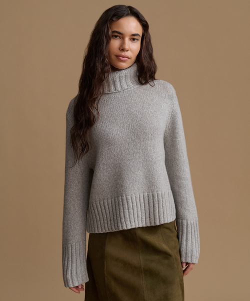 A woman wearing a light gray ribbed turtleneck sweater and an olive green skirt, standing against a solid tan studio background.