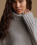 A close-up of a woman wearing a chunky, ribbed knit turtleneck sweater in a light gray color, with her hand adjusting the collar.