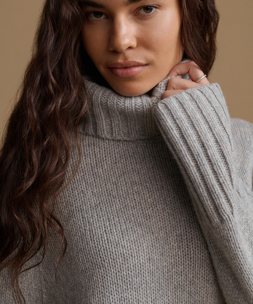 A close-up of a woman wearing a chunky, ribbed knit turtleneck sweater in a light gray color, with her hand adjusting the collar.