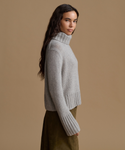 A woman wearing a light gray ribbed turtleneck sweater and an olive green skirt, shown in profile against a solid beige studio background.
