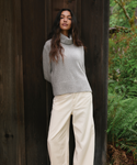 A woman wearing a light gray cowl-neck knit sweater and cream wide-leg trousers, standing against a dark wooden wall with green foliage visible on the right.