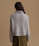 A woman shown from the back wearing a light gray ribbed knit turtleneck sweater and an olive green skirt, set against a solid tan studio background.