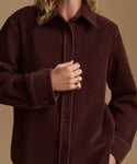 A close-up of a person wearing a dark brown corduroy button-up shirt jacket against a solid beige background.