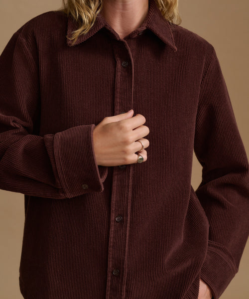 A close-up of a person wearing a dark brown corduroy button-up shirt jacket against a solid beige background.