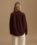 A woman wearing a dark brown corduroy button-up shirt and cream trousers, standing against a solid beige studio background.