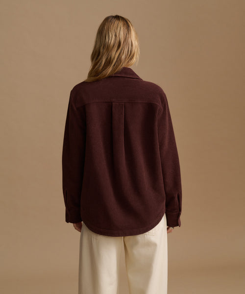 A woman wearing a dark brown corduroy button-up shirt and cream trousers, standing against a solid beige studio background.