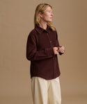 A woman wearing a dark brown corduroy button-up shirt and cream trousers, standing against a solid beige studio background.