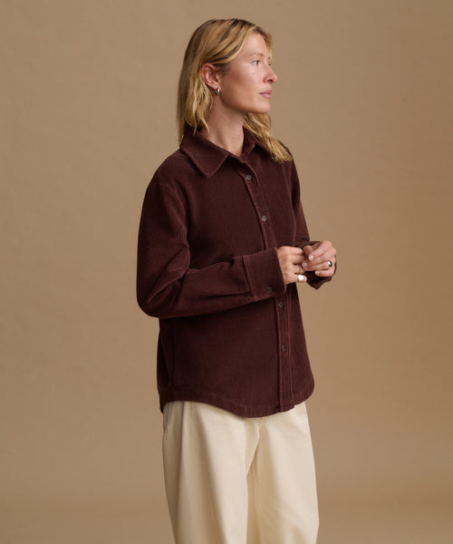 A woman wearing a dark brown corduroy button-up shirt and cream trousers, standing against a solid beige studio background.