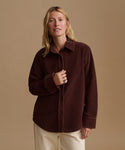 A woman wearing a dark brown corduroy button-up shirt and cream trousers, standing against a solid beige studio background.