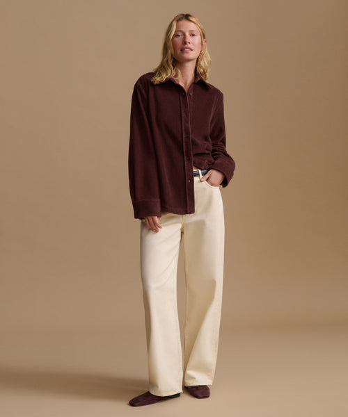 A woman wearing a dark brown corduroy button-up shirt and cream trousers, standing against a solid beige studio background.