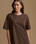 A woman wearing a brown short-sleeve crewneck t-shirt, photographed against a solid warm beige studio background.