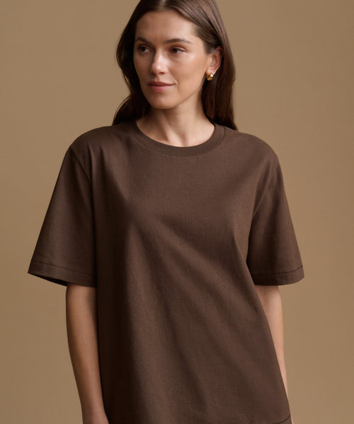 A woman wearing a brown short-sleeve crewneck t-shirt, photographed against a solid warm beige studio background.