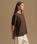 A woman wearing an oversized brown short-sleeve t-shirt and cream-colored pants, shown in profile against a solid beige studio background.