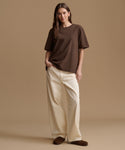 A woman modeling an oversized brown short-sleeve t-shirt and cream wide-leg pants against a solid beige studio background.