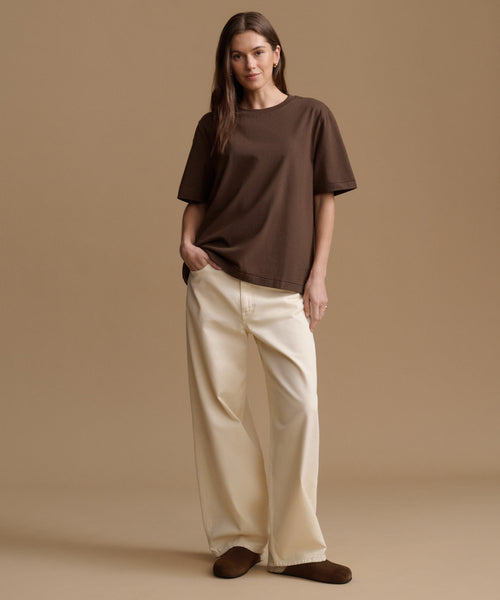 A woman modeling an oversized brown short-sleeve t-shirt and cream wide-leg pants against a solid beige studio background.