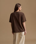 A woman shown from the back wearing an oversized dark brown short-sleeve t-shirt and cream-colored pants, standing against a solid beige studio background.
