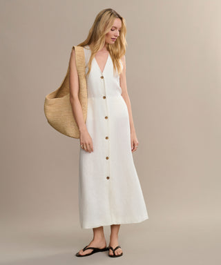 A woman wears the sleeveless white Callan Dress with black sandals, carrying a large woven straw bag over her shoulder, standing against a plain beige background.