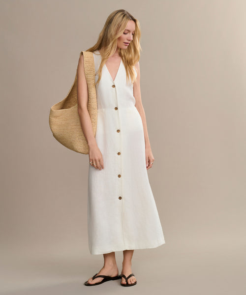 A woman wears the sleeveless white Callan Dress with black sandals, carrying a large woven straw bag over her shoulder, standing against a plain beige background.