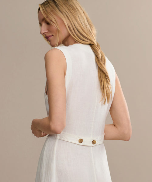 A woman with long blonde hair in a loose braid is shown from the back wearing the sleeveless white Callan Dress, which features a fabric belt with two buttons at the waist, set against a plain beige background.