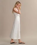 A woman with long blonde hair wears the sleeveless, ankle-length Callan Dress in white and black sandals, standing in profile against a plain beige background. The Callan Dress is a timeless staple for classic elegance.