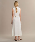 A woman with long blonde hair in a braid stands with her back to the camera, wearing the sleeveless, ankle-length white Callan Dress featuring button details at the waist and black sandals against a plain beige background.