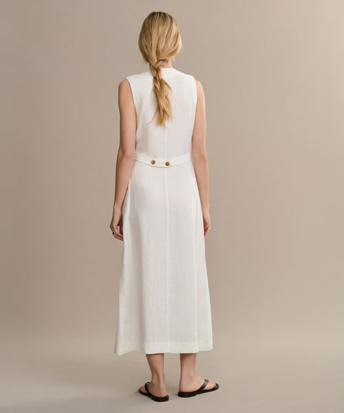 A woman with long blonde hair in a braid stands with her back to the camera, wearing the sleeveless, ankle-length white Callan Dress featuring button details at the waist and black sandals against a plain beige background.