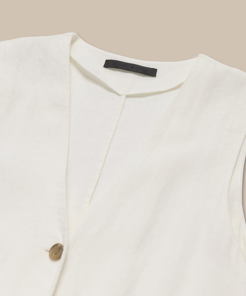 A close-up of the Callan Dress, a sleeveless white linen staple with a V-neckline and single brown button, displayed flat on a beige background with a black label inside the collar.