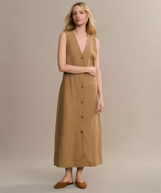 A woman with loose blonde hair stands against a beige background, wearing the Callan Dress—a sleeveless tan V-neck vest dress—paired with brown flats, calmly looking at the camera to showcase this closet staple.