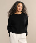 A woman with long, curly gray hair wears the Cameron Crewneck sweater with white pants, standing against a plain beige background and smiling slightly at the camera.