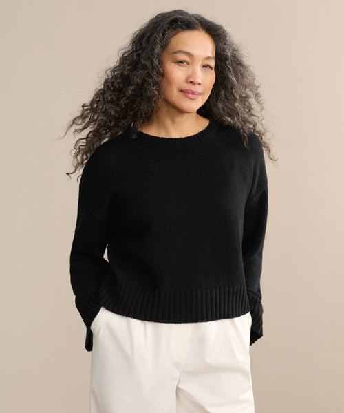 A woman with long, curly gray hair wears the Cameron Crewneck sweater with white pants, standing against a plain beige background and smiling slightly at the camera.