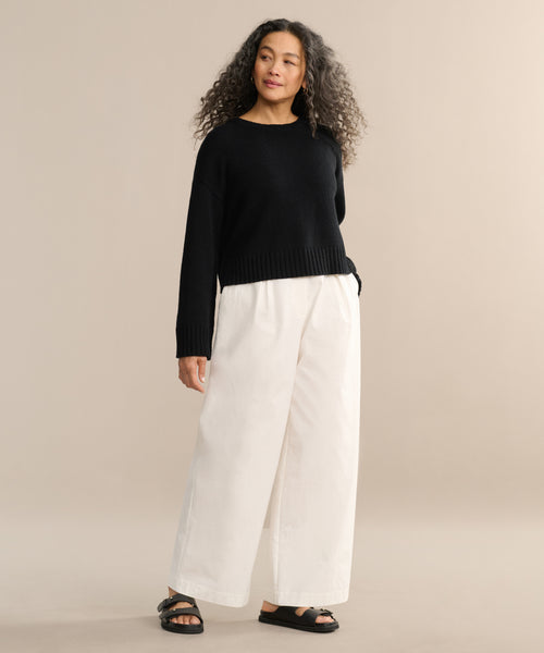 A woman with long curly hair models the Cameron Crewneck sweater in black, paired with wide-leg white pants and black sandals. She stands against a neutral beige background, gazing to the side with a relaxed expression.