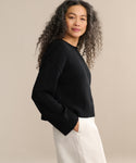 A woman with long, curly hair wears the Cameron Crewneck sweater in black and white pants, standing confidently with one hand in her pocket against a plain beige background.