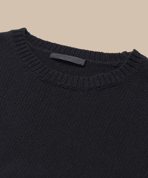 Close-up of the Cameron Crewneck, a black lightweight sweater with a ribbed neckline, laid flat on a beige background. A small black label is visible inside the collar.