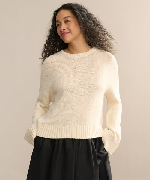 A woman with long, curly hair smiles slightly against a neutral background, wearing the cream-colored Cameron Crewneck sweater and a black skirt, her hands in her pockets.